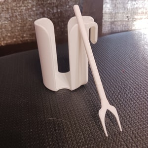 Può includere: Un supporto in plastica bianca con un design a forma di U e un piccolo gancio curvo. Una forchetta di plastica bianca a due rebbi è appoggiata accanto al supporto. Gli oggetti sono su una superficie scura e strutturata.