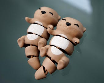 May include: Two brown otter figurines with articulated limbs and white accents on their bellies. The otters have black eyes and are smiling. The figurines are made of a matte material and are posed in a playful arrangement.