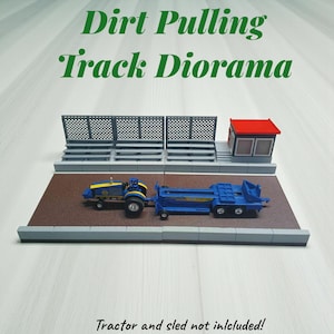 Puede incluir: Diorama en miniatura de una pista de arrastre de tierra con un tractor azul y amarillo tirando de un trineo azul. La pista presenta vallas grises, una cabina con techo rojo y una superficie de pista marrón. Se muestra el texto "Dirt Pulling Track Diorama".