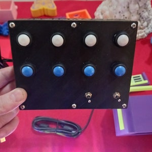 May include: A black box with eight buttons, four white and four blue. The buttons are arranged in two rows of four. There are two small switches on the bottom of the box.