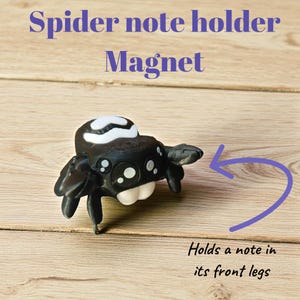 May include: A black and white spider-shaped magnet note holder. The spider has large, friendly eyes and holds a note in its front legs. The text "Spider note holder Magnet" is at the top of the image.