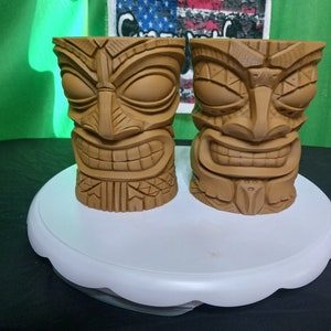 Può includere: Due tazze tiki marroni con volti intagliati e motivi geometrici. Le tazze sono su un piedistallo bianco.