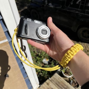 May include: Black HP Photosmart M447 digital camera with a yellow wrist strap. The camera is held by a yellow paracord bracelet. The camera has a silver lens and flash.
