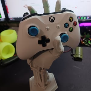 May include: A white Xbox controller is held by a 3D printed robotic hand. The hand is attached to a hexagonal base. The controller has blue buttons and a black directional pad.