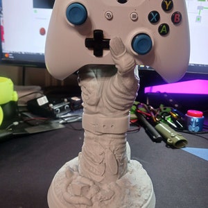 May include: A white 3D printed figurine of a person holding a white Xbox controller. The figurine is standing on a round base.