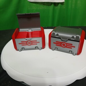 May include: Two red and gray toy chests with a retro design. One chest is open, revealing the interior. The chests have a circular emblem and horizontal lines. The chests are on a white surface.