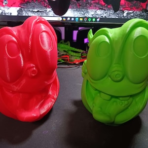 May include: Two 3D printed figurines with cartoonish faces. One is red and the other is green.