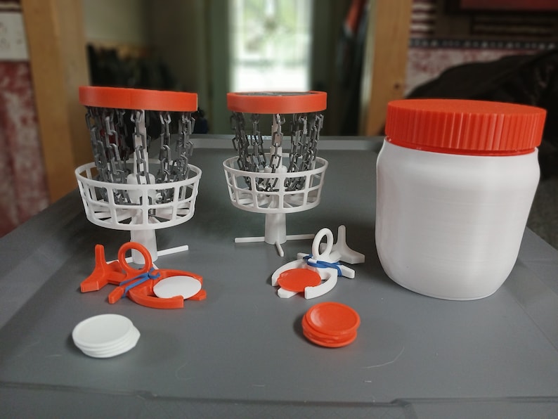 May include: A set of miniature disc golf baskets with orange rims and white bases, along with orange discs and a white container with an orange lid.