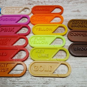 May include: A selection of brightly coloured keychains, each labelled with a different colour name, such as 'Fuchsia', 'Candy Red', and 'Key Lime'. The keyrings are made of a 3D-printed material and are displayed on a light wood-grain surface.