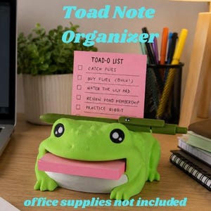 May include: A bright green frog-shaped note organizer featuring a wide mouth holding a stack of pink sticky notes. A pink notepad with the text TOAD-O LIST and five tasks is inserted into the top, with a green pen resting horizontally across it.