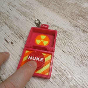 以下が含まれることがあります： 黄色い放射能記号と「NUKE」と書かれた赤いボタンが付いた赤いプラスチック製のオモチャのブリーフケースです。
