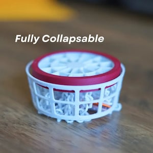 May include: A white collapsible basketball hoop with a red rim. The hoop is fully collapsed and is sitting on a wooden surface. The text "Fully Collapsible" is written above the hoop.