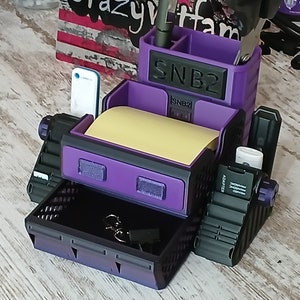 May include: A purple and black desk organizer with a drawer, a compartment for pens and scissors, and a sticky note holder. The organizer has the text "SNB2" on the front.