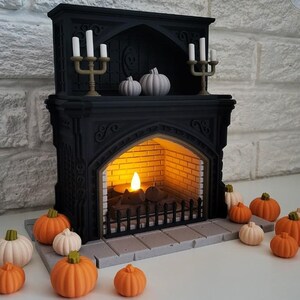 Puede incluir: Una chimenea negra con una vela encendida en su interior y un candelabro decorativo dorado con velas blancas en la parte superior. Hay varias calabazas peque&ntilde;as de color naranja y blanco delante de la chimenea.