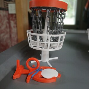May include: A miniature disc golf basket with a white chain and a red top. A red disc golf disc is lying on the ground next to the basket.