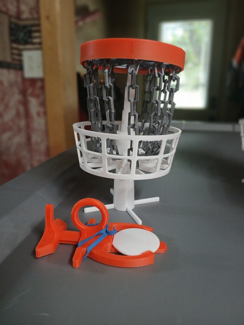 May include: A miniature disc golf basket with a white chain and a red top. A red disc golf disc is lying on the ground next to the basket.