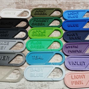 May include: A selection of 3D-printed carabiner clips in various colours. Each clip is labelled with a colour name, such as white, black, army green, forest green, light blue, blue, navy blue, mystic purple, violet, and light pink.