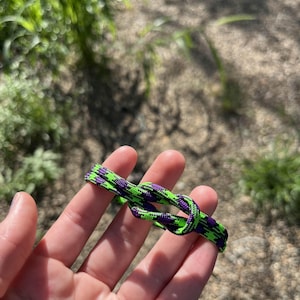 May include: A neon green and purple paracord bracelet is held in a hand. The bracelet is knotted and features a repeating pattern of the two colors. The background is blurred, showing greenery and a path.