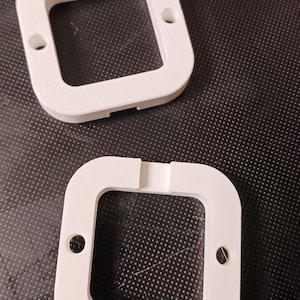 May include: Two white plastic square-shaped frames with a smaller square opening in the centre. The frames are designed to hold a square object in place.