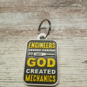 May include: A gray and white keychain with the text "Engineers needed heroes so God created mechanics". The text is in yellow and white. A small screwdriver is pictured above the word "God".