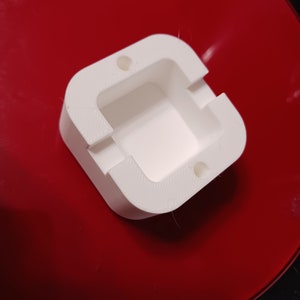 May include: White plastic mold with a square opening and two small holes on the top. The mold is sitting on a red surface.