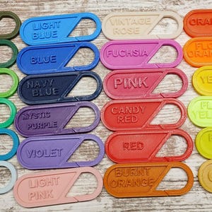 May include: A selection of plastic carabiner clips in various colours, each labelled with a colour name. Shades include white, black, light grey, space grey, silver, army green, forest green, apple green, teal, baby blue, light blue, blue, navy blue, mystic purple, violet, light pink, vintage rose, fuchsia, pink, candy red, red, burnt orange, orange, flor orange, yellow, flor yellow, key lime, gold, coyote brown, copper, and brown.