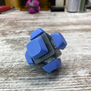 Puede incluir: Un pequeño juguete antiestrés azul y gris con seis lados. Cada lado tiene una superficie azul texturizada.