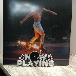 May include: A black and white vinyl record stand with the words "NOW PLAYING" and musical notes. A black vinyl record is on the stand. The background image is a man on roller skates with flames coming from the wheels.