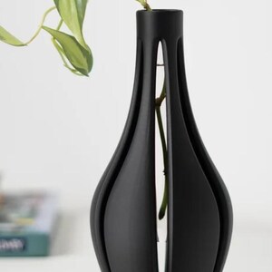 May include: A black ceramic vase with a unique, modern design. The vase has a narrow neck and a wide, rounded base. It features a series of vertical cuts that create a geometric pattern. The vase is empty, but it is ready to hold flowers or other decorative elements.