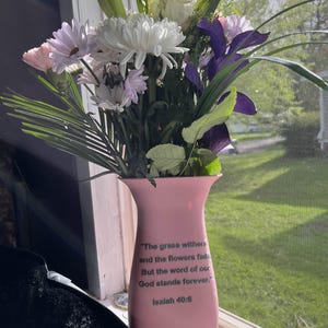 May include: A pink vase with a floral arrangement of white, purple, and pink flowers. The vase has a quote from Isaiah 40:8 printed on it: "The grass withers and the flowers fade, But the word of our God stands forever."