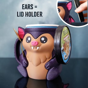 May include: A novelty bat-shaped mug in purple and tan, with a unique lid-holding design. The mug has large eyes, a pink nose, and small fangs. The bat's ears are designed to hold the lid. The text "EARS = LID HOLDER" is visible.