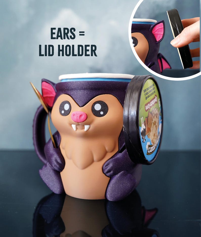 May include: A novelty bat-shaped mug in purple and tan, with a unique lid-holding design. The mug has large eyes, a pink nose, and small fangs. The bat's ears are designed to hold the lid. The text "EARS = LID HOLDER" is visible.
