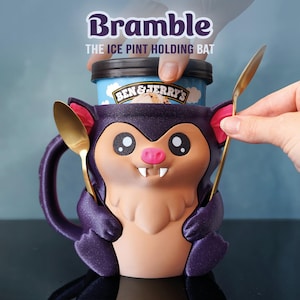 May include: A novelty ice cream pint holder in the shape of a purple bat, with a cartoon face and two gold spoons. The holder is designed to hold a pint of ice cream. The text "Bramble" and "THE ICE PINT HOLDING BAT" are displayed on the image.