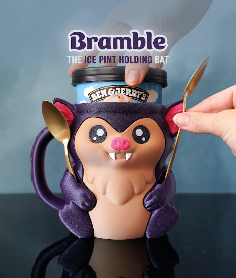 May include: A novelty ice cream pint holder in the shape of a purple bat, with a cartoon face and two gold spoons. The holder is designed to hold a pint of ice cream. The text "Bramble" and "THE ICE PINT HOLDING BAT" are displayed on the image.