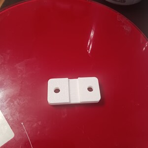Könnte beinhalten: Weißes, 3D-gedrucktes Kunststoffteil mit zwei Löchern und einem Schlitz in der Mitte. Das Teil liegt auf einer roten Oberfläche.