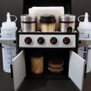 Może przedstawiać: Miniaturowy czarny i szary grill z czterema palnikami, dwiema białymi butelkami do ściskania, dwoma stalowymi potrząsaczami i pojemnikiem na wykałaczki. Grill ma białą pokrywę z białym papierowym ręcznikiem w środku. Grill ma dwie białe drzwi, które otwierają się, ukazując schowek z większą ilością wykałaczek i pojemnikiem na przyprawy.