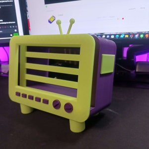 Puede incluir: Un televisor retro amarillo y morado con un estante en el interior. El televisor tiene una parte trasera morada y una parte delantera amarilla con una perilla morada en la parte inferior derecha.