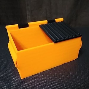 May include: A small orange plastic dumpster with a black lid. The lid has a ribbed texture and is slightly ajar.