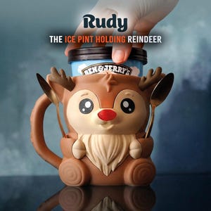 May include: A brown reindeer-shaped ice cream pint holder with the text "Rudy THE ICE PINT HOLDING REINDEER". The holder has a red nose, large eyes, and holds a pint of ice cream. Two golden spoons are placed on the sides.