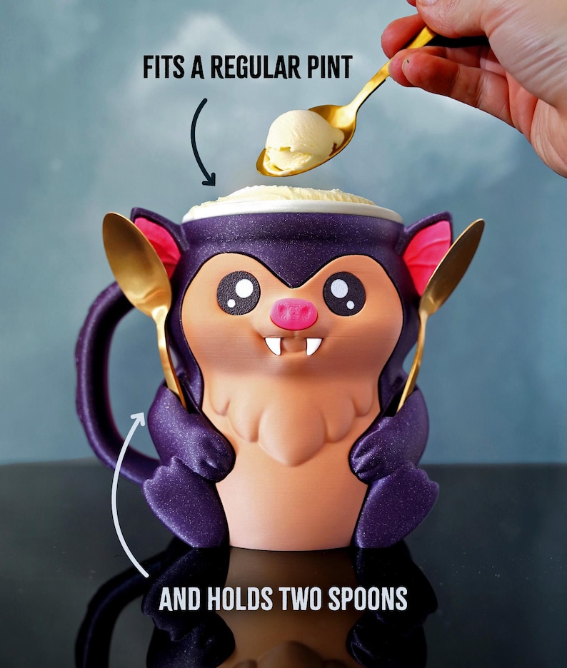 May include: A novelty purple bat-shaped mug with a pink nose and large eyes, holding two gold spoons. The mug is filled with ice cream, and a spoon is scooping more ice cream into the mug. Text on the image reads: FITS A REGULAR PINT and AND HOLDS TWO SPOONS.
