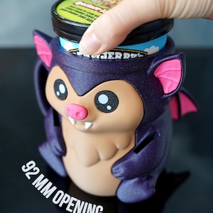 May include: A purple bat-shaped ice cream container holder with pink accents. The holder has a 9.2 cm opening and is designed to hold a pint of ice cream. The bat has large eyes, a pink nose, and small fangs. A hand is placing an ice cream container into the holder.