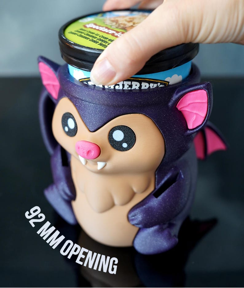 May include: A purple bat-shaped ice cream container holder with pink accents. The holder has a 9.2 cm opening and is designed to hold a pint of ice cream. The bat has large eyes, a pink nose, and small fangs. A hand is placing an ice cream container into the holder.