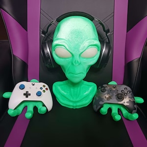 May include: A green alien bust with headphones on, holding two video game controllers in its hands. The controllers are a white Xbox controller and a camouflage Xbox controller.