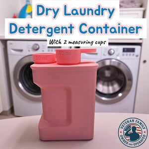 Może przedstawiać: Różowy pojemnik na suchy detergent do prania z dwiema miarkami, jedną 30 ml i drugą 50 ml. Pojemnik ma napis "Dry Laundry Detergent Container" i "With 2 measuring cups". Pojemnik stoi na stole przed pralką.