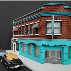 May include: A miniature model of a brick building with a turquoise awning and shop front. The building has a sign that reads "Sodas" and "Ice Cream". There are two cars parked in front of the building.