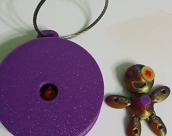 May include: A purple keychain with a round, textured base and a metal ring. A small, colorful, 3D printed figure of a person with a smiling face is attached to the ring.