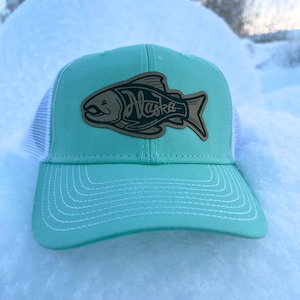 May include: A light green and white trucker hat with a black and white fish graphic and the word "Alaska" on it.