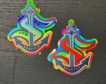 May include: Two iridescent stickers featuring a stylized anchor with the Space Needle and the word "Seattle". The anchor is outlined in black and the Space Needle is red on one sticker and blue on the other.