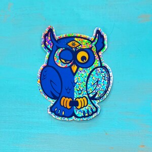 May include: A blue and silver glitter sticker of an owl with a third eye on its forehead. The owl is winking and has a serious expression.