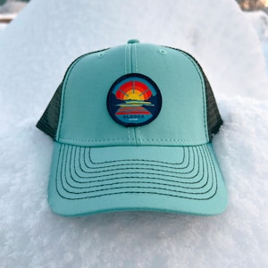 May include: A teal blue trucker hat with a black mesh back. The hat has a patch on the front with a sunset scene and the word "ALASKA" in white text.